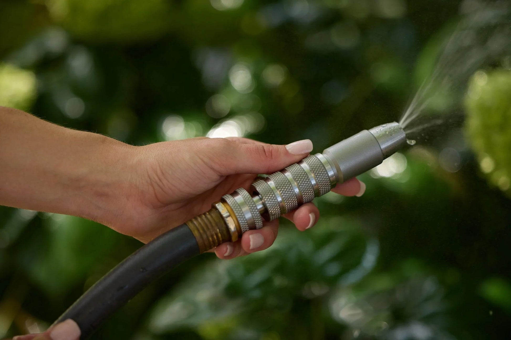 Hand holding stainless steel spray nozzle attached to garden hose, spraying water outdoors